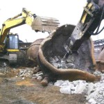 One excavator stabilizes a ladle so that another can hammer out the hardened slag.